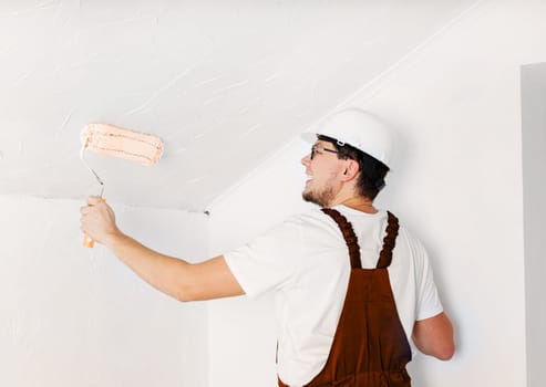 Color of the year 2024: Peach Fuzz. Repair, home improvement and renovation concept. young man in overalls and hard hat painting wall into white color with a roller