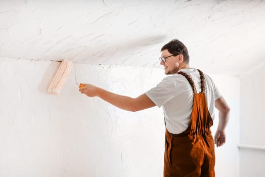 Color of the year 2024: Peach Fuzz. Repair, home improvement and renovation concept. young man in overalls painting wall into color with a rolle