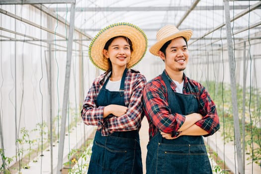 Happiness blooms in tomato hydroponic farm with Asian couple. Smiling farmers crossed arms carrying quality vegetables. Portrait of successful husband and wife in the greenhouse. Farming joy.