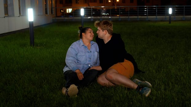 Happy romantic couple sitting on the green grass near building and street lamps. Media. Young man and woman on green lawn