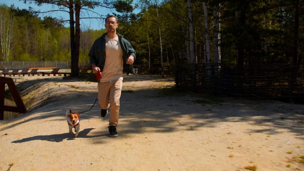 Happy man and his little cute puppy running along the green small lake. Stock footage. Man jogging with jack russell