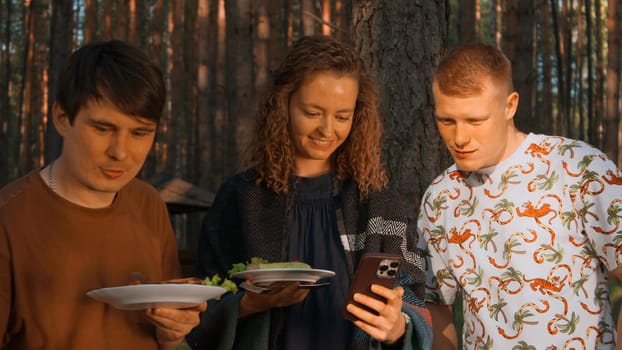 Group of friends eating barbecue and laughing fun by the grill. Stock footage. Girl showing something funny in her smartphone