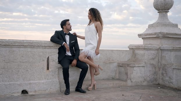 A young couple of models. Action. A woman in a short white dress walks along the pier with a man in a suit putting her long legs on him. High quality 4k footage
