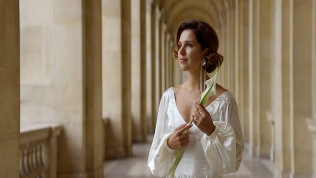 Beautiful young romantic woman in a long white dress standing near ancient columns. Action. Beautiful woman holding a flower in her hand