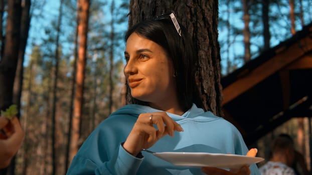 Beautiful brunette woman standing by the tree and eating. Stock footage. Woman taking food from plate, smiling on the background of forest and sky