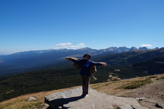 joyful smiling elderly woman with raised hands stands on top of mountain.