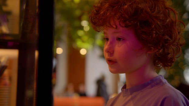 Red haired cute boy on the background of the interior with green leaves. Stock footage. Curly hair of a little boy