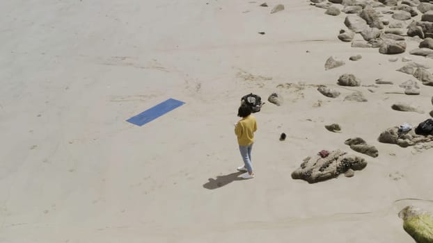 Aerial view of a woman wearing yellow sweater and blue jeans standing on the sandy sea shore near her yoga mat. Female standing on the sand near the stones.