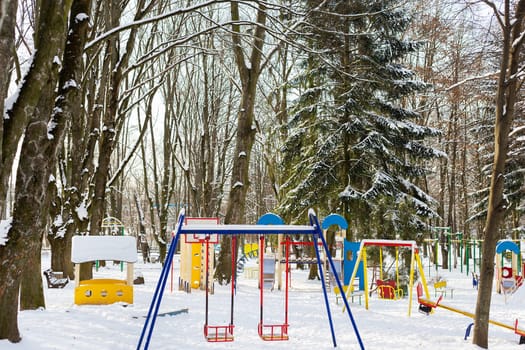 A quiet winter playground covered in snow, with colorful equipment and surrounded by bare evergreen trees