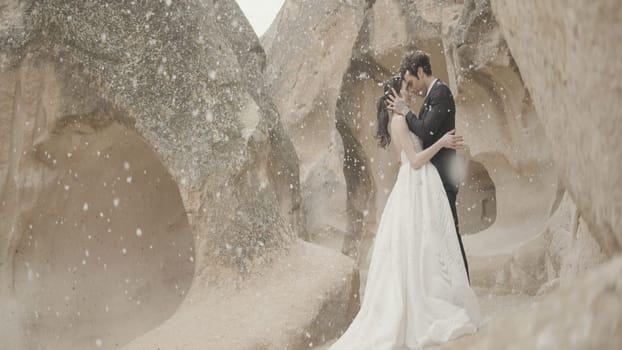 Newlyweds at stones with snow. Action. Beautiful couple of newlyweds embracing on background of natural stone wall. Newlyweds hug at stone wall in snowfall.