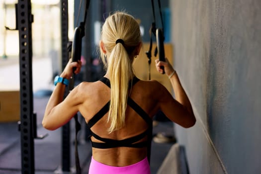 Back view of strong fit woman with ponytail wearing sportswear and doing exercise with gymnastics rings during fitness workout in modern gym
