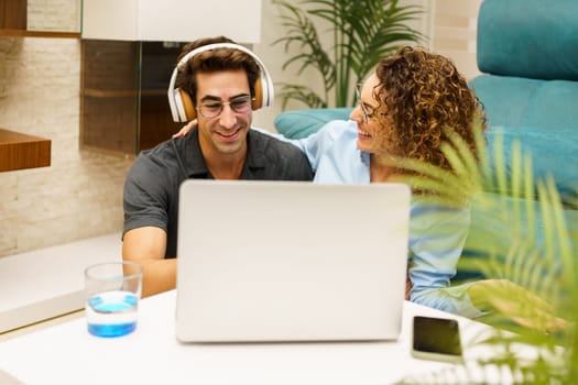 Cheerful young woman sitting near man wearing wireless headphones and using laptop at home