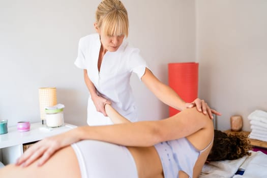 Concentrated crop masseuse looking down, while standing doing physiotherapy in osteopathy clinic to young slim anonymous female shoulder lying on bed and pulling hand in bright daylight