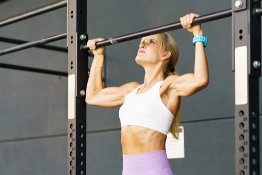 Fit young muscular female in activewear hanging on metal horizontal bar while exercising during fitness workout at modern gym club against gray wall