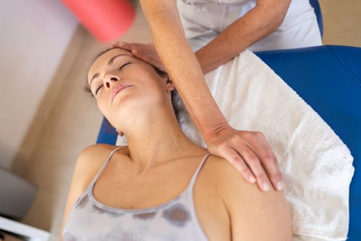 From above crop osteopath stretching neck of patient lying on white towel in bed with eyes closed during physiotherapy treatment in clinic