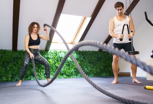 Full body of young curly haired female athlete in sportswear exercising with battle ropes during workout with fitness buddy in modern gym