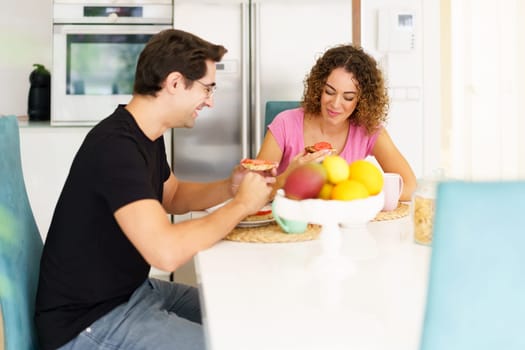 Happy adult couple in conversation and looking down while sitting at dining table in kitchen with fruits in bowl, and eating breakfast in daylight at home