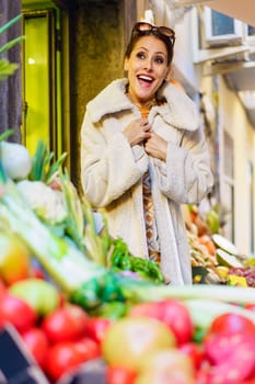 Low angle of excited young woman in warm coat with hands by chest standing near stall with bunch of fresh vegetables, and ripe fruits while smiling and looking at camera