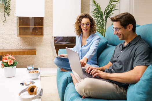 Positive young husband and wife in smart casual clothes and eyeglasses sitting on sofa and browsing laptop while working on remote project in bright