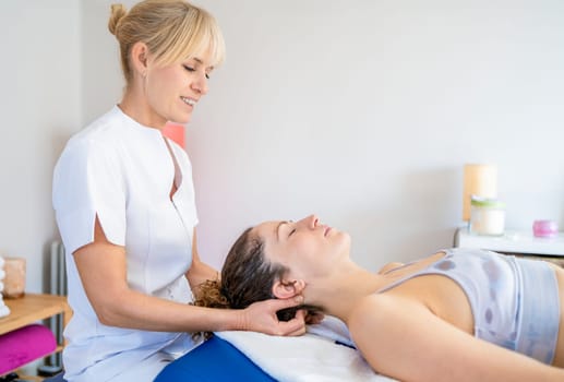 Side view of blonde hair osteopath in white uniform sitting and massaging neck of patient lying in bed and closing eyes during physiotherapy session