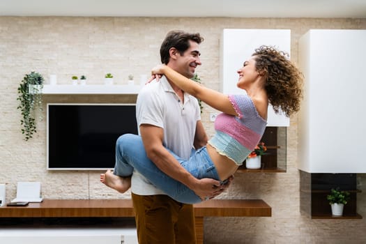 Side view of cheerful young romantic couple embracing while enjoying time against decorated background with potted plants and TV in modern apartment