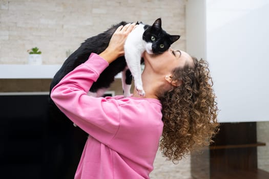 Side view of crop anonymous female in casual cloth with curly brown hair lifting and kissing adorable cat, with black and white fur while in room at home