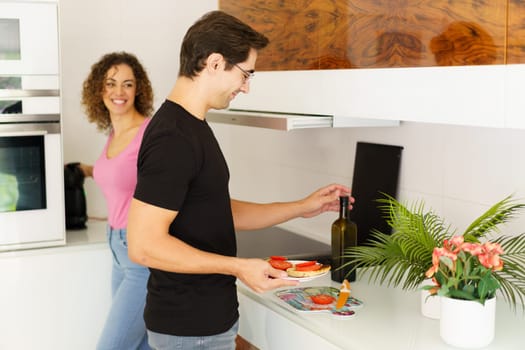 Side view of happy adult male looking down while standing with smiling conversing female at counter and carrying healthy salad, while opening wine bottle near flower pot in kitchen in daylight