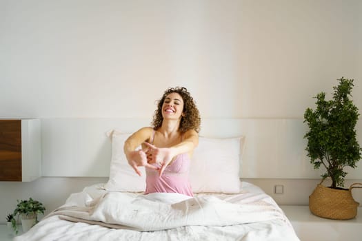 Smiling adult female with curly hair and eyes closed while sitting on cozy bed in daylight and stretching reverse clasped hands, in front with interlocked fingers near green plant