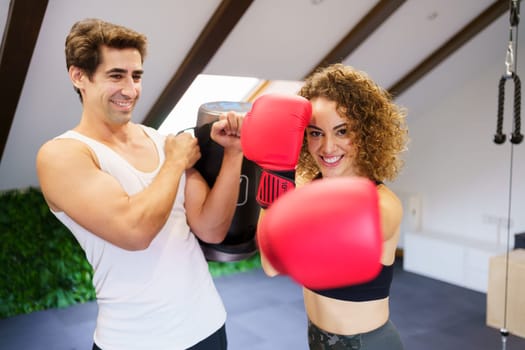 Smiling curly haired strong female fighter in red boxing gloves punching air during training with personal instructor holding bag in gym