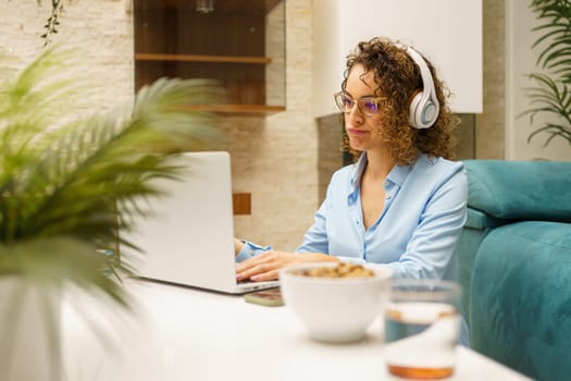 Tired young curly haired female freelancer in casual clothes and eyeglasses with headphones sitting at breakfast table, and browsing laptop while working on remote project in living room