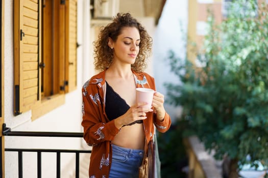 Young stylish female with curly hair in fashionable outfit smiling and holding cup while standing on balcony of modern building against blurred green trees