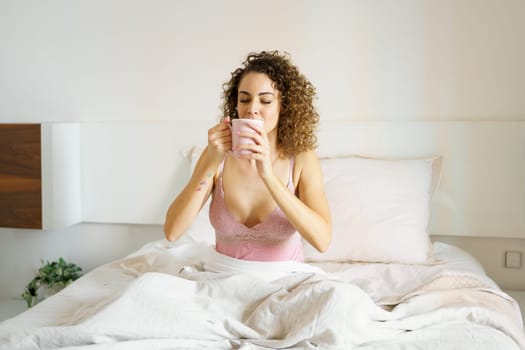 Young woman in nightwear drinking fresh hot coffee while sitting with eyes closed in bed at home