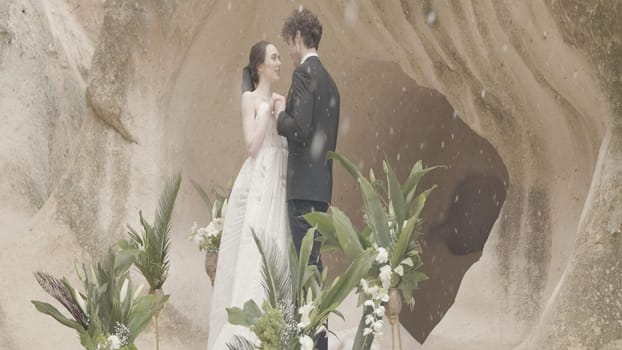 Newlyweds at stones with snow. Action. Beautiful couple of newlyweds embracing on background of natural stone wall. Newlyweds hug at stone wall in snowfall.