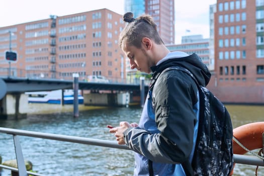 Fashionable young male with backpack using smartphone outdoors in city. Hipster with trendy hairstyle beard with phone in hands mobile applications for tourism travel communication entertainment study