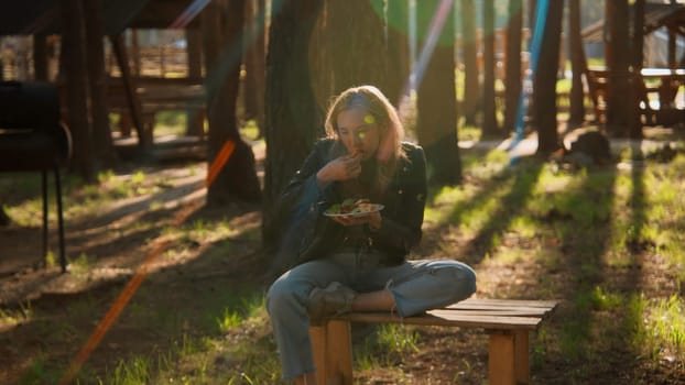 Young woman sitting on bench and eating barbecue and vegetables. Stock footage. Woman with barbecue plate and shining sun through the trees