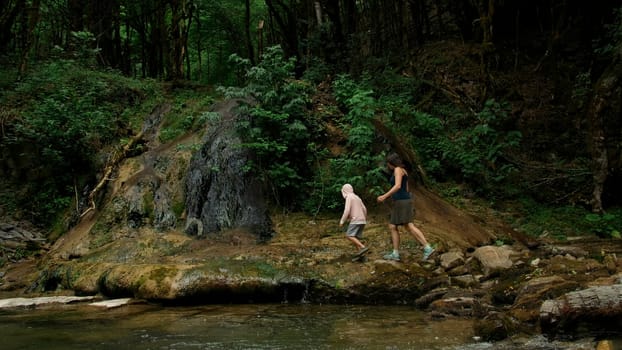 Young boy and his mother walking along the mountainous river near the cliff. Creative. Green forest and stones