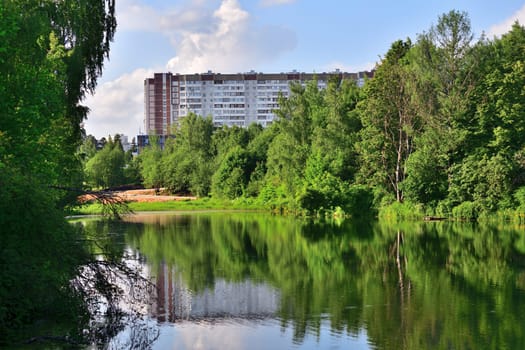 Summer landscape with a pond surrounded by trees and a multi-story building in the background