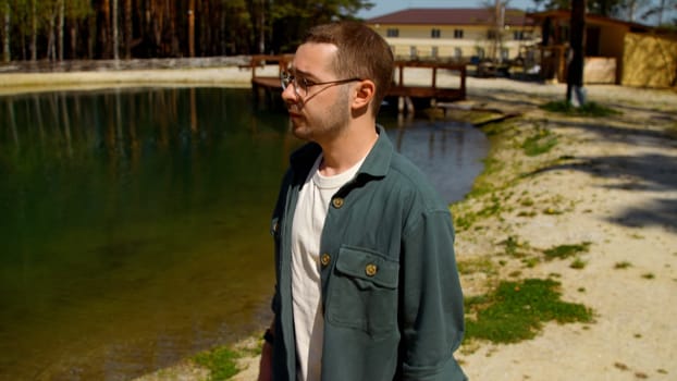 Young depressed man in glasses looking unhappy. Stock footage. Man walking by the small green pond and countryside houses