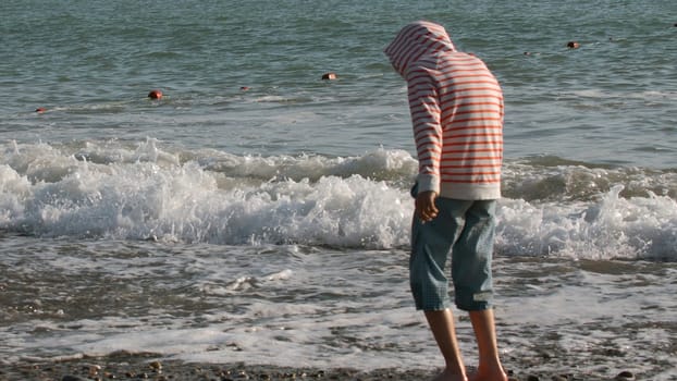 Young boy standing barefoot in the water on the pebble shore. Creative. Rear view of a boy exploring sea shore
