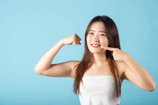 Dental health strong powerful. Beautiful woman point fingers to smiling mouth and raises arms and shows biceps studio shot isolated on blue background, Asian young woman pointing to strong white teeth