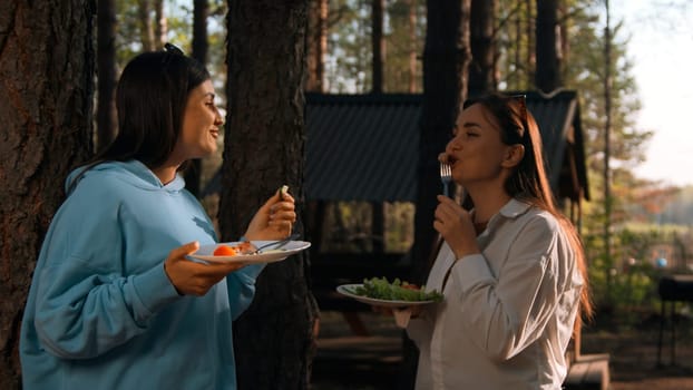 Female friends standing by the tree with plates and eating food while talking and smiling. Stock footage. Camping with friends in forest