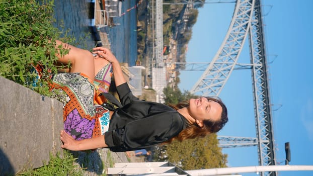 Vertical video with beautiful woman on embankment. Action. Attractive woman is sitting on edge of embankment on sunny summer day. Stylish woman is resting on city embankment with view of bridge.