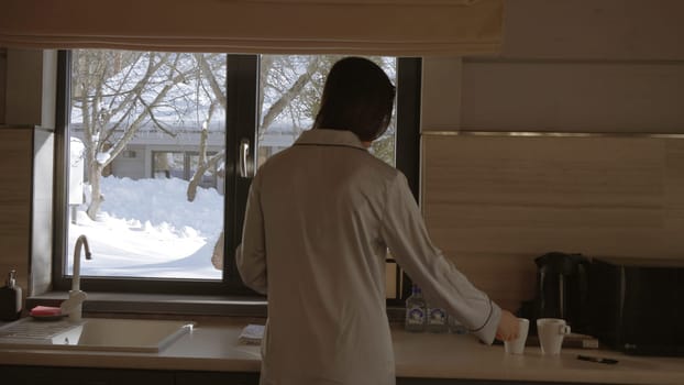 Rear view of a woman making hot beverage in front of the window and winter outside. Action. Cozy kitchen and a girl making morning tea