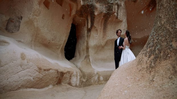 Stylish beautiful bride and groom under falling snow near rocks or caves. Action. Wedding video shooting in cappadocia