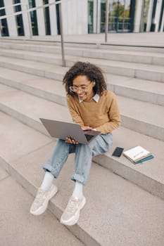 Smart young woman student in eyeglasses is using pc laptop sitting outdoors on building background