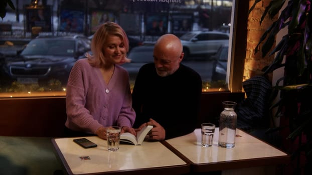 Happy middle aged mature couple talking together at the restaurant in the evening. Stock footage. Smiling loving senior old family having fun, enjoying on date in cafe with a book on the table