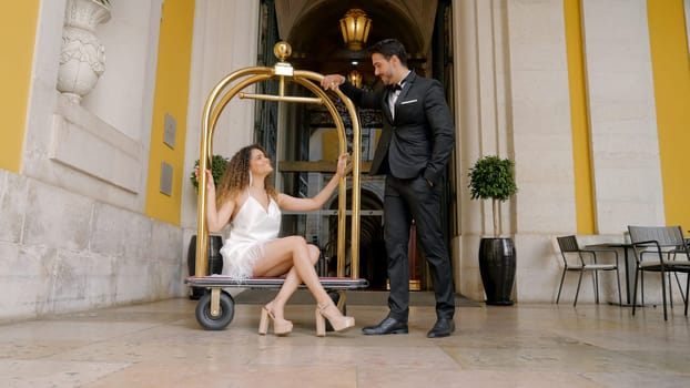 Elegant couple at entrance to hotel. Action. Elegant woman sits on European luggage trolley in hotels. Beautiful luxury couple at expensive hotel.