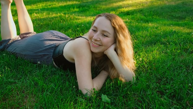 Beautiful young woman lying on grass in park in summer. Concept. Beautiful blonde woman is lying on grass and talking on sunny day. Attractive young woman is resting on summer green grass.