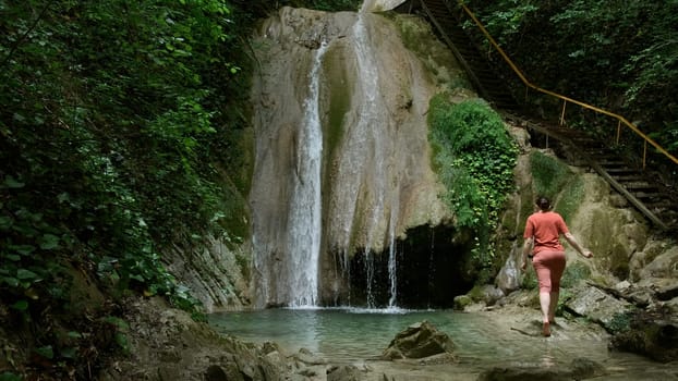 Woman near waterfall located in summer green forest. Creative. Woman walking barefoot towards the waterfall near the picturesque touristic wooden stairs