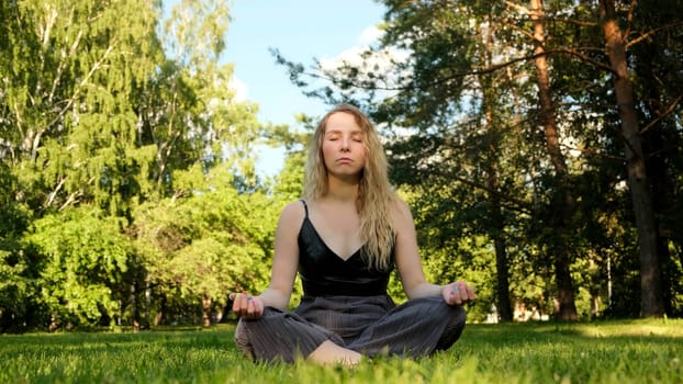 Young beautiful relaxed blonde woman sitting on green grass and meditating. Concept. Unity with nature, meditation in a city park